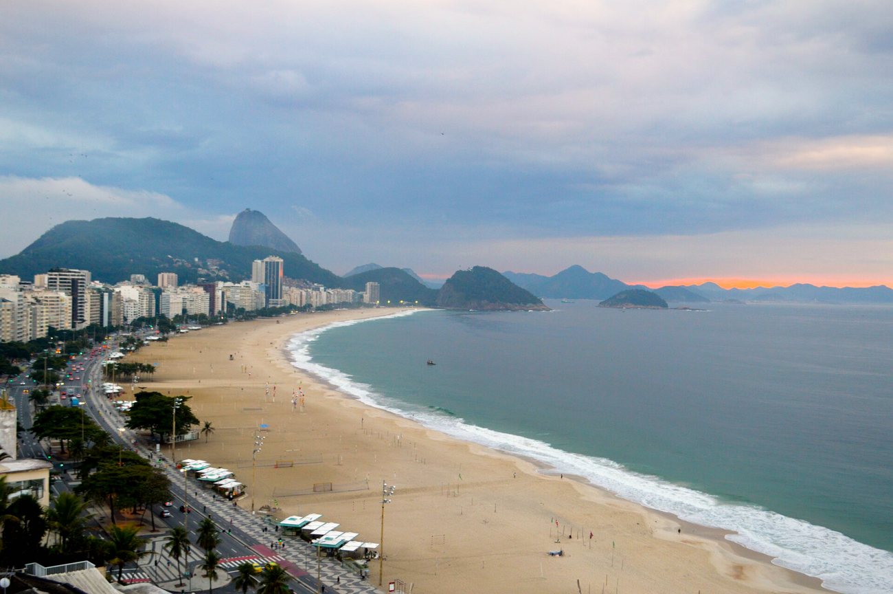 Copacabana Beach at dusk seen from above, with the curve of the shoreline, beach kiosks and apartment towers below Sugarloaf Mountain